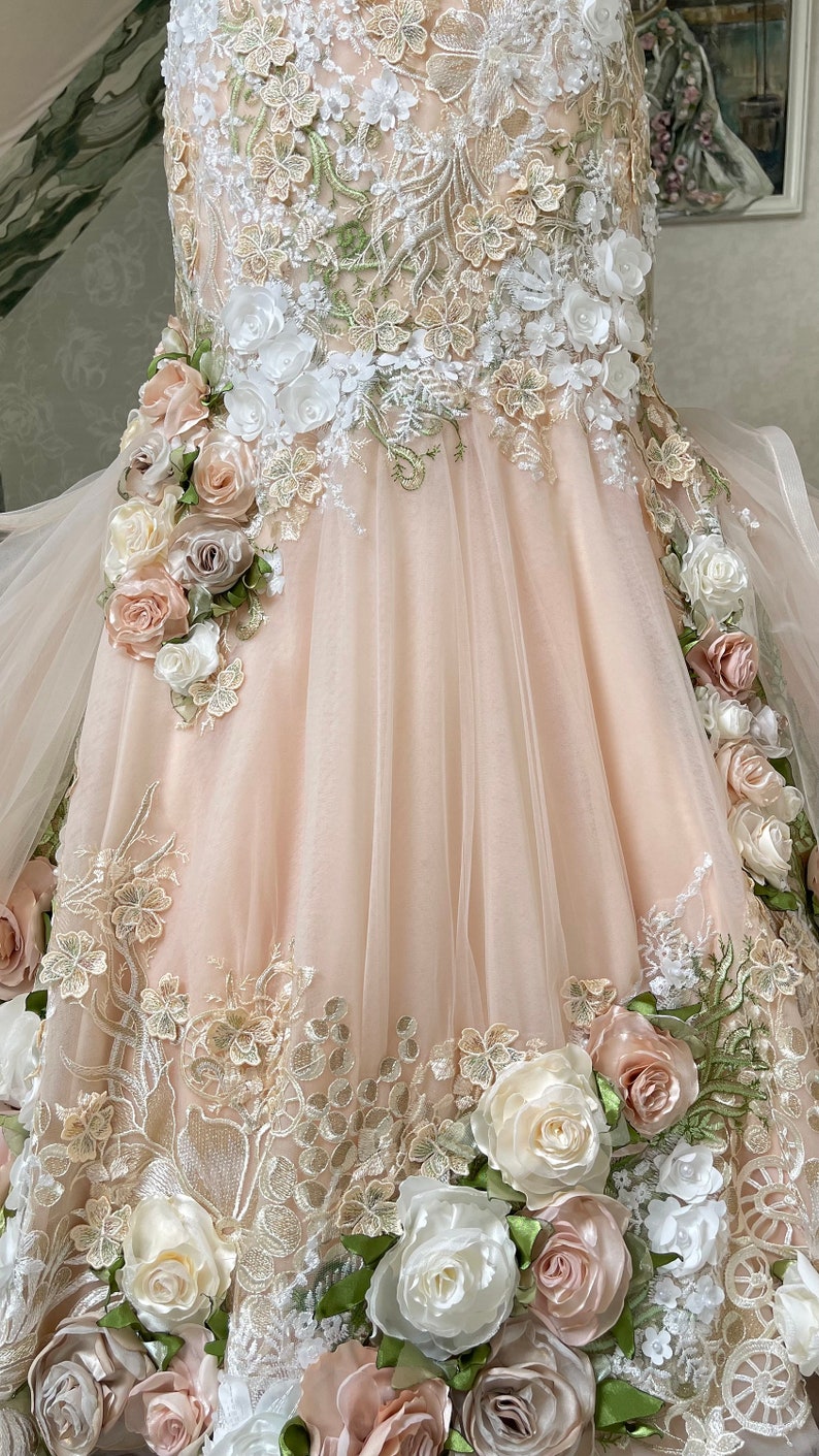 Puede incluir: Un vestido de tul rosa claro con intrincados bordados florales y aplicaciones florales 3D. El vestido presenta una variedad de flores en tonos de blanco, rosa y verde.