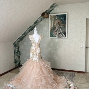 Puede incluir: Un vestido de novia de tul rosa claro con un corpi&ntilde;o ajustado y una falda completa. El corpi&ntilde;o est&aacute; adornado con aplicaciones florales blancas y doradas. La falda tiene varias capas de tul y est&aacute; adornada con un ribete floral.