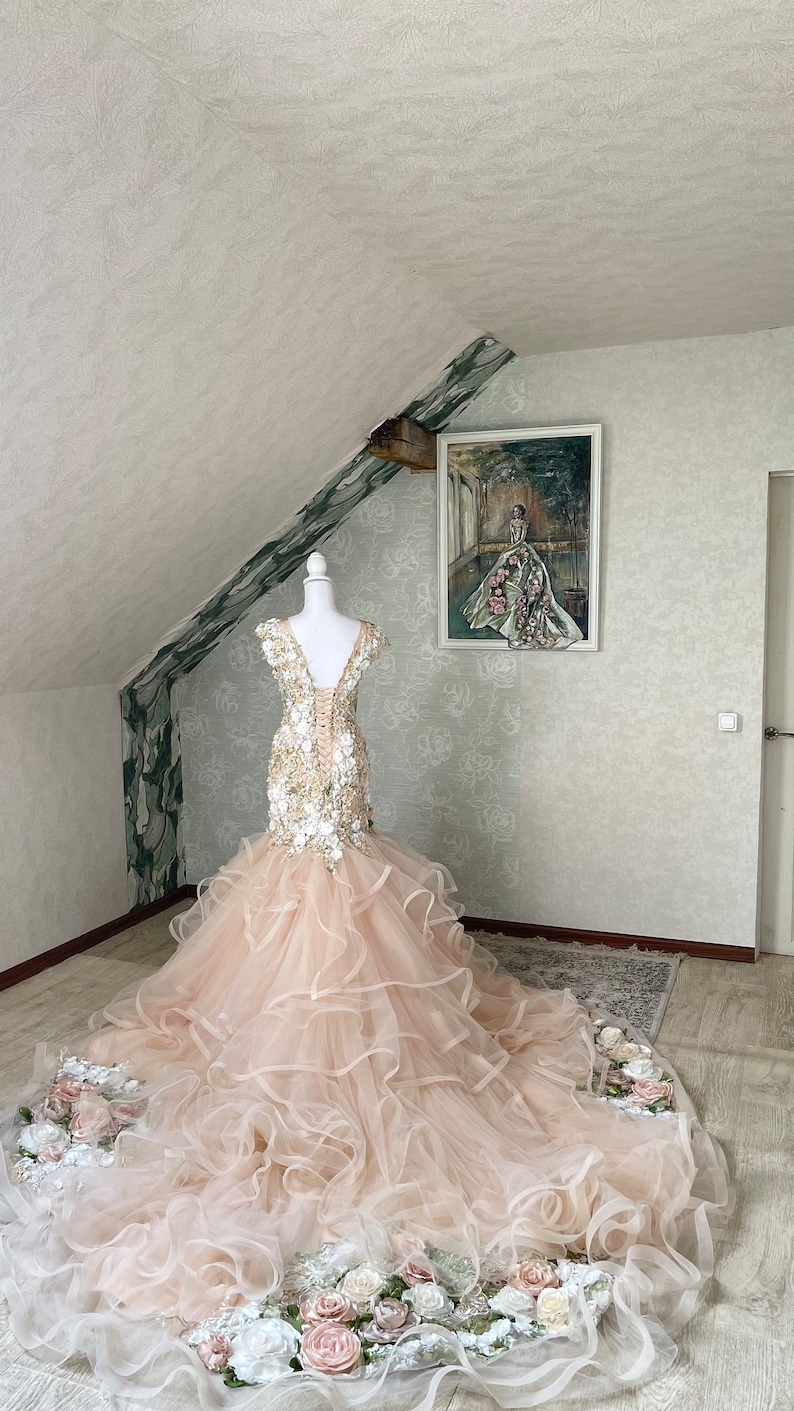 Puede incluir: Un vestido de novia de tul rosa claro con un corpi&ntilde;o ajustado y una falda completa. El corpi&ntilde;o est&aacute; adornado con aplicaciones florales blancas y doradas. La falda tiene varias capas de tul y est&aacute; adornada con un ribete floral.
