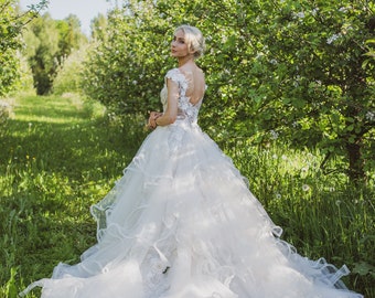 May include: A bride in a white wedding dress with lace details stands in a field of green grass and trees. The dress has a full skirt and a fitted bodice. The bride is looking over her shoulder.