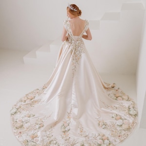 May include: A cream-colored wedding gown with floral embroidery and a long train. The dress features a low back and cap sleeves. The train is adorned with 3D floral appliques in shades of pink, cream, and green. A tiara is worn on the head.