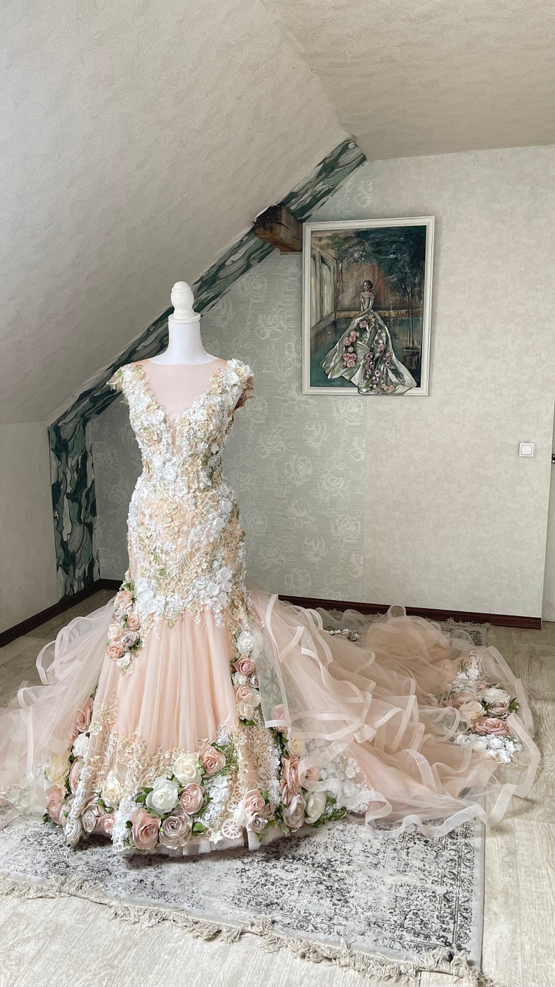 Puede incluir: Un vestido de novia rosa empolvado con un corpi&ntilde;o ajustado y una falda fluida. El vestido est&aacute; adornado con delicados apliques florales y una larga cola.