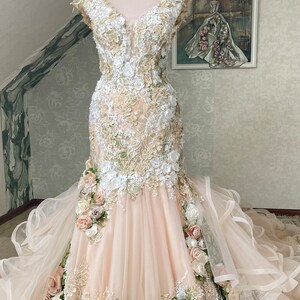 Puede incluir: Un vestido de novia de tul rosa claro con un corpi&ntilde;o ajustado y una falda fluida. El vestido est&aacute; adornado con encaje delicado y aplicaciones florales 3D en tonos de blanco, rosa claro y verde.