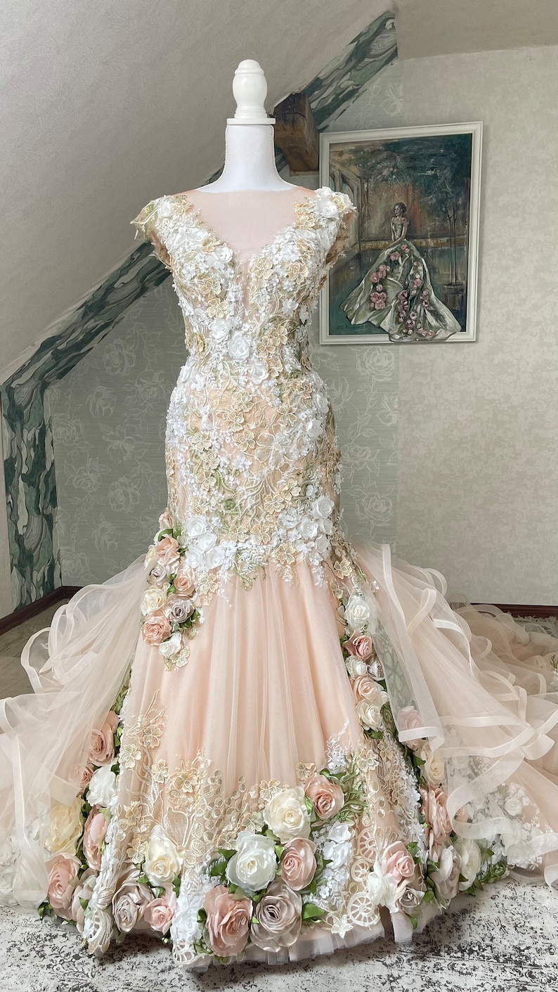 Puede incluir: Un vestido de novia de tul rosa claro con un corpi&ntilde;o ajustado y una falda fluida. El vestido est&aacute; adornado con encaje delicado y aplicaciones florales 3D en tonos de blanco, rosa claro y verde.