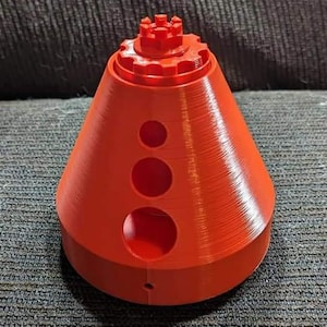 May include: A bright orange 3D printed cone-shaped object with a circular opening at the top and three smaller circular openings on the side. The top of the object has a gear-like design.