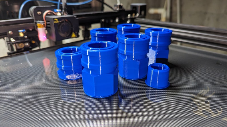 May include: A collection of bright blue 3D-printed plastic connectors. The connectors have a cylindrical shape with a hexagonal base and a grooved design. The objects are arranged on a dark surface, with a 3D printer visible in the background.