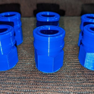 May include: Six bright blue, 3D-printed quick-connect fittings are arranged on a brown surface. Each fitting has a cylindrical top and a hexagonal base. The fittings are made of plastic.
