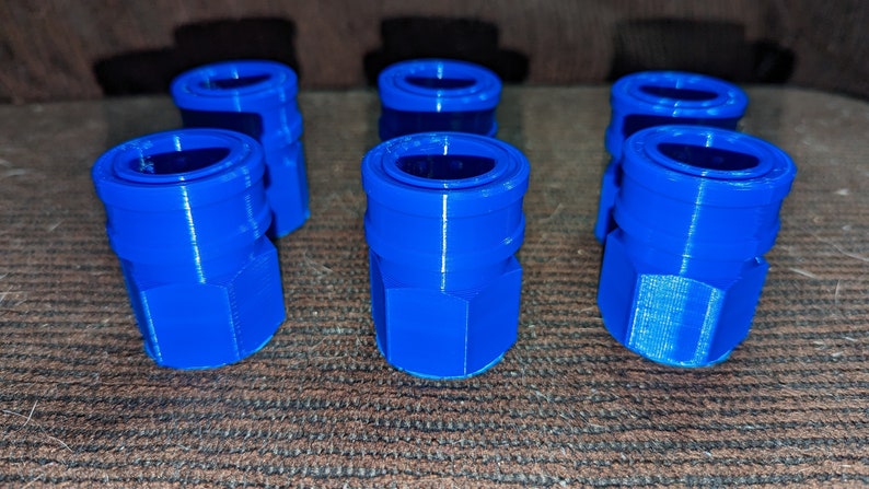 May include: Six bright blue, 3D-printed quick-connect fittings are arranged on a brown surface. Each fitting has a cylindrical top and a hexagonal base. The fittings are made of plastic.