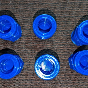 May include: Six bright blue, 3D-printed plastic objects are arranged on a brown, textured surface. Each object has a hexagonal base and a circular top with a central opening. The objects are evenly spaced.