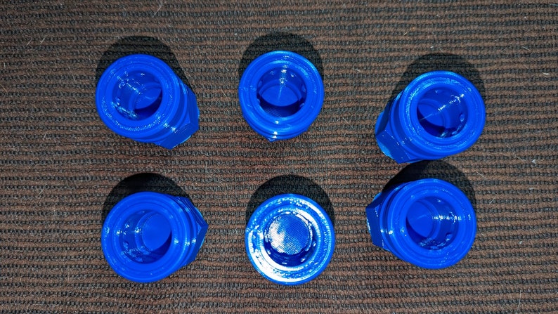 May include: Six bright blue, 3D-printed plastic objects are arranged on a brown, textured surface. Each object has a hexagonal base and a circular top with a central opening. The objects are evenly spaced.