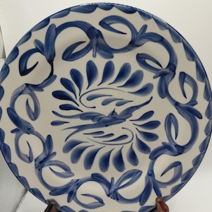 May include: A round ceramic plate featuring a hand-painted blue floral and leaf pattern on a white background. The center of the plate displays a stylized bird motif surrounded by radiating brushstrokes, with a decorative border of vines.