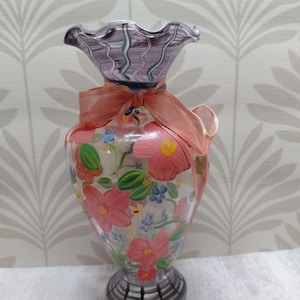 May include: A clear glass vase with a ruffled, purple-tinted rim and a matching purple-tinted base featuring black vertical lines. The body is decorated with hand-painted pink flowers, green leaves, and small blue blossoms, tied with a peach ribbon.