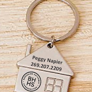 May include: Silver house-shaped keychain with a key ring. The keychain has the text "Peggy Napier 269.207.2209" and a Berkshire Hathaway HomeServices logo.