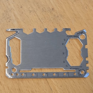 Puede incluir: Una tarjeta multiherramienta plateada con varias funciones, incluyendo un abrebotellas, un cortador de alambre y una regla. La tarjeta tiene un transportador, un orificio para llavero y un abridor de gafas. La regla está marcada en cm.