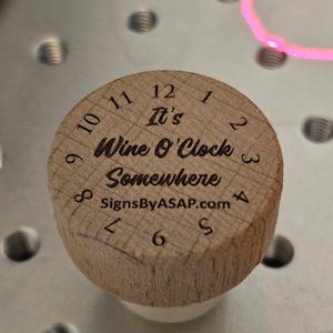 May include: A wooden bottle stopper with the text "It's Wine O'Clock Somewhere" and the website "SignsByASAP.com" engraved on it. The stopper has a clock face with numbers 1 through 12.