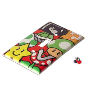 Pode incluir: Uma capa decorativa para interruptor de luz com um design colorido de personagens de desenhos animados sobre um fundo vermelho. A capa inclui uma estrela amarela sorridente, um cogumelo verde e outros gr&aacute;ficos relacionados a jogos. Inclui parafusos de montagem.
