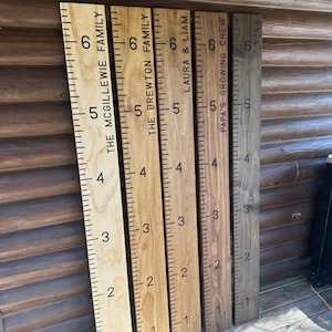 May include: Wooden growth charts with family names and height markings. The charts are in various wood tones, including natural, golden oak, and dark walnut. Each chart has a ruler-style measurement scale in inches, with family names and labels.