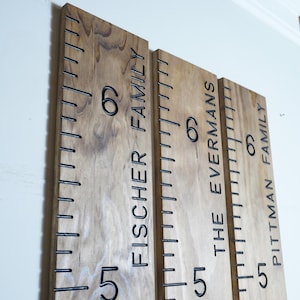 May include: Three wooden growth charts with black measurements and family names: Fischer Family, The Evermans, and Pittman Family. Each chart has numbers 5 and 6, with inch markings. The wood has a natural grain.