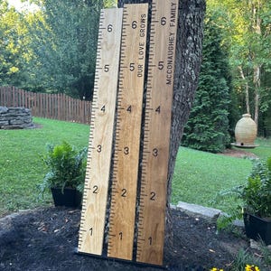May include: Three wooden growth charts leaning against a tree. The charts are marked with numbers from 1 to 6, with inch markings. One chart reads "OUR LOVE GROWS", and another reads "MCCONAUGHEY FAMILY".