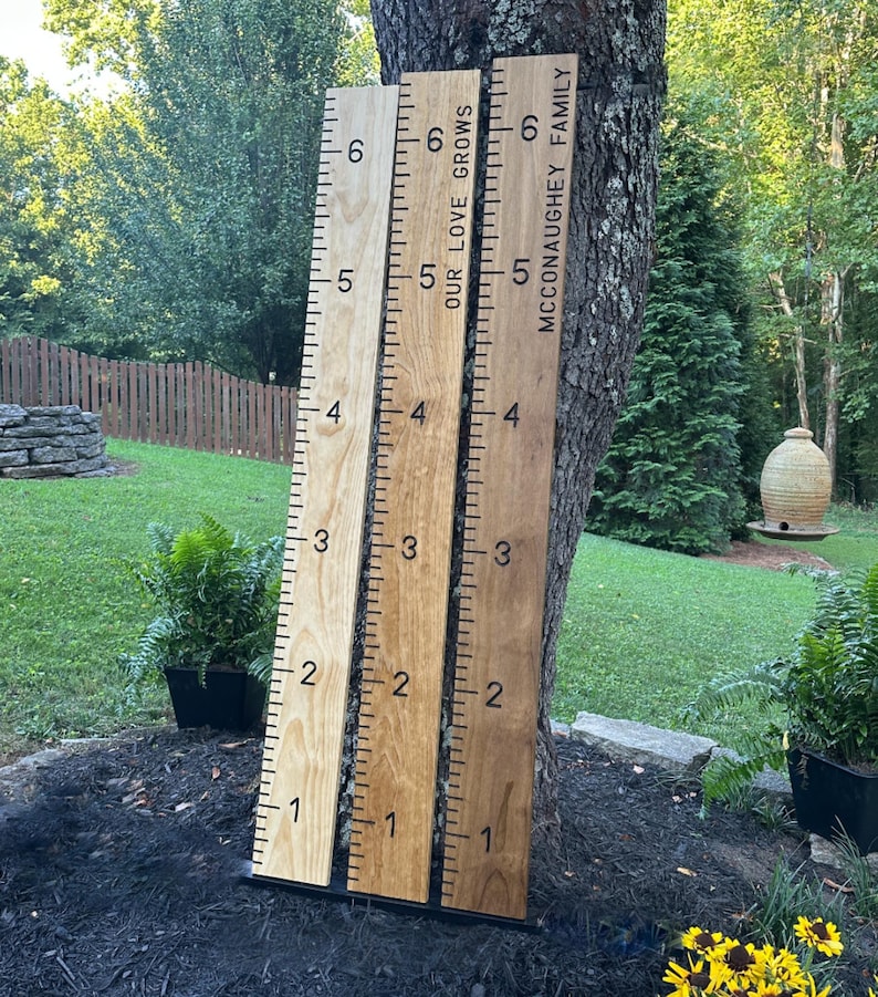 May include: Three wooden growth charts leaning against a tree. The charts are marked with numbers from 1 to 6, with inch markings. One chart reads "OUR LOVE GROWS", and another reads "MCCONAUGHEY FAMILY".