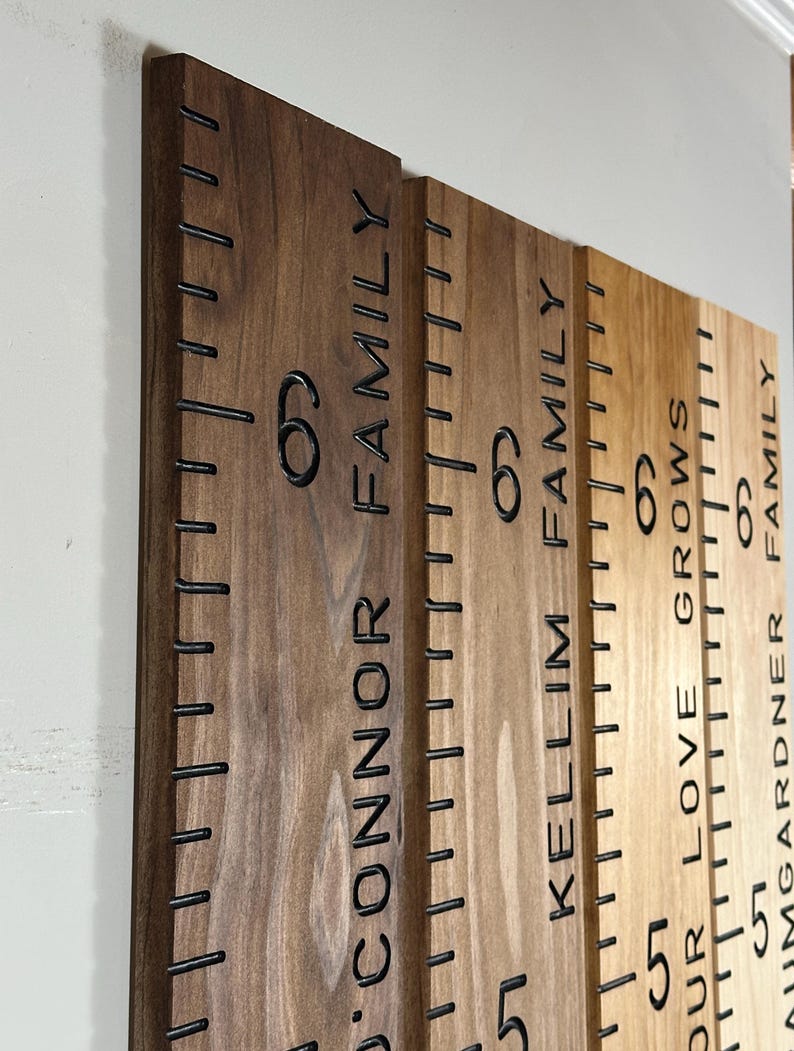 May include: Wooden growth chart with engraved height markings and family names. The chart features black markings and numbers, with the text "O'Connor Family", "Kelim Family", and "Our Love Grows". The wood has a natural finish.
