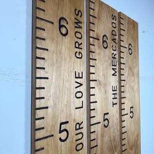 May include: Three wooden growth charts with black markings and numbers. The text "OUR LOVE GROWS" and "THE MERCADOS" are vertically printed on the boards. The charts are mounted on a light blue wall.