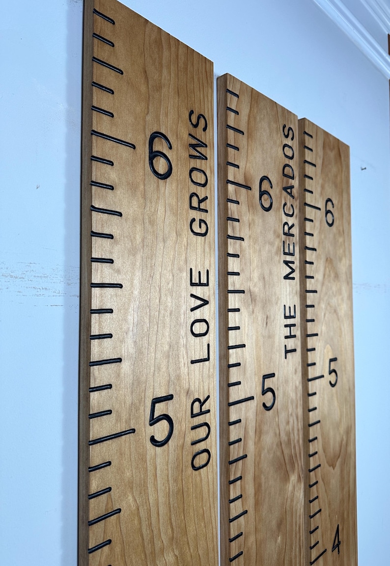 May include: Three wooden growth charts with black markings and numbers. The text "OUR LOVE GROWS" and "THE MERCADOS" are vertically printed on the boards. The charts are mounted on a light blue wall.