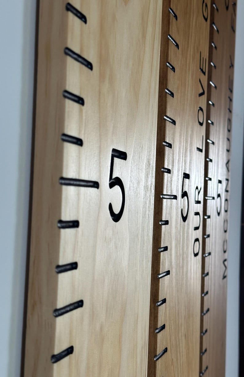 May include: Wooden growth chart with black markings and the number 5 in a bold font. The chart has black metal staples marking the height increments. The words "OUR LOVE" and a name are visible on the side of the chart.