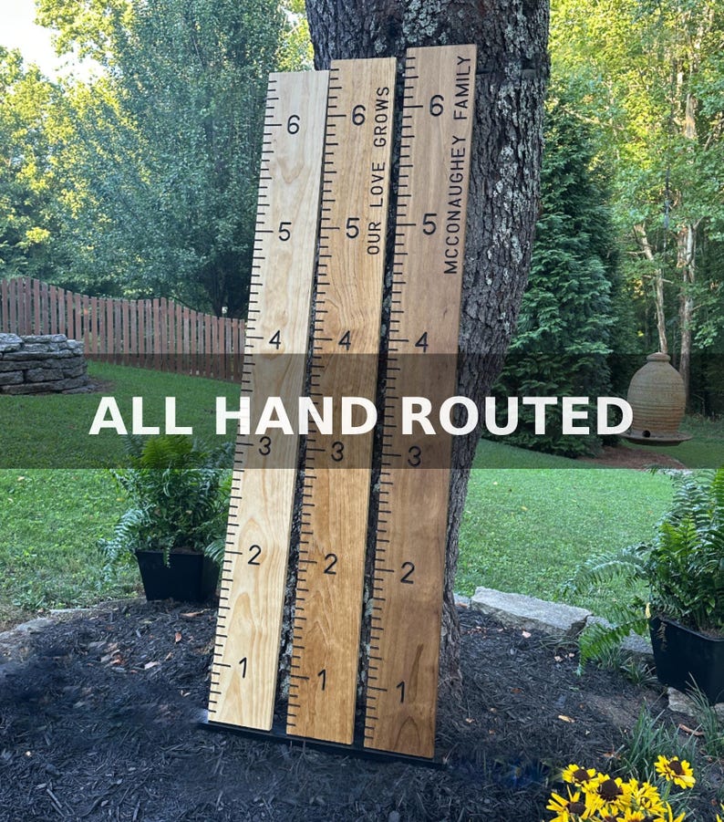 May include: Three wooden growth charts with ruler markings and the text "OUR LOVE GROWS" and "MCCONAUGHEY FAMILY". The charts are in varying shades of brown and are leaning against a tree. The text "ALL HAND ROUTED" is at the bottom.