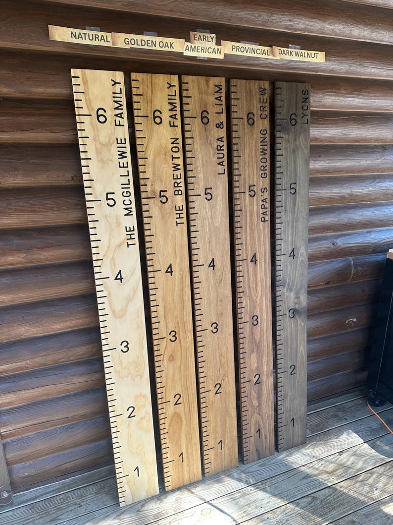 May include: Wooden growth charts with family names and height markings. The charts are in various wood tones, including natural, golden oak, and dark walnut. Each chart has a ruler-style measurement scale in inches, with family names and labels.