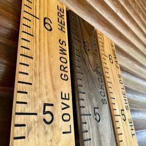 May include: Wooden growth charts with black measurement markings and engraved text. The charts are in various wood tones, with phrases like "LOVE GROWS HERE" and family names. Each chart has numbers indicating height.