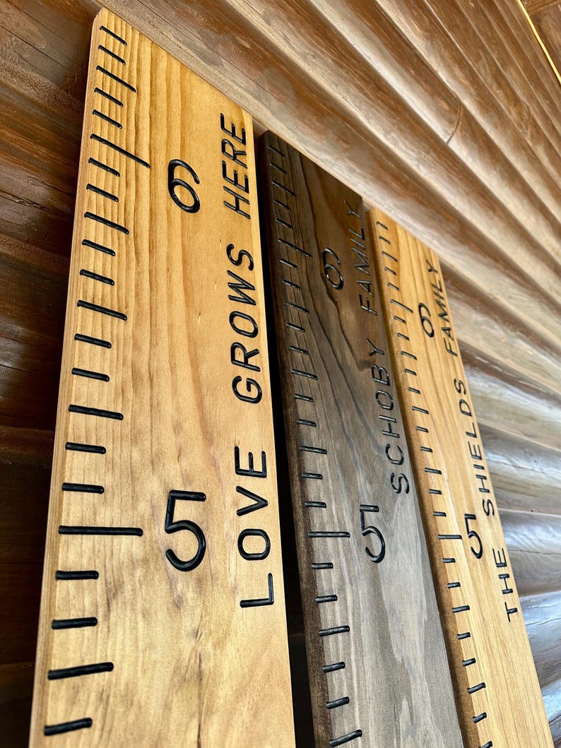 May include: Wooden growth charts with black measurement markings and engraved text. The charts are in various wood tones, with phrases like "LOVE GROWS HERE" and family names. Each chart has numbers indicating height.