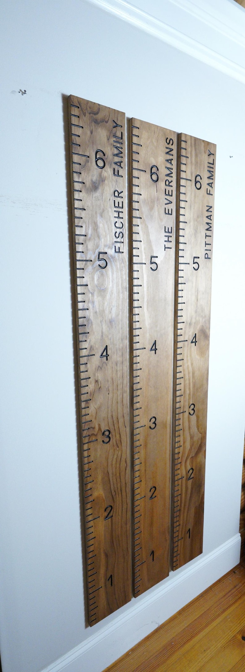 May include: Three wooden growth charts mounted on a white wall. Each chart is engraved with a family name and numbered markings from 1 to 6 feet. The charts are made of light brown wood.