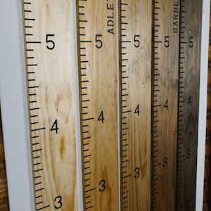 May include: Wooden growth charts in various finishes: Natural, Golden Oak, Early American, Provincial, and Dark Walnut. Each chart is marked with black numbers and lines, measuring up to 6 feet. Some charts include names.