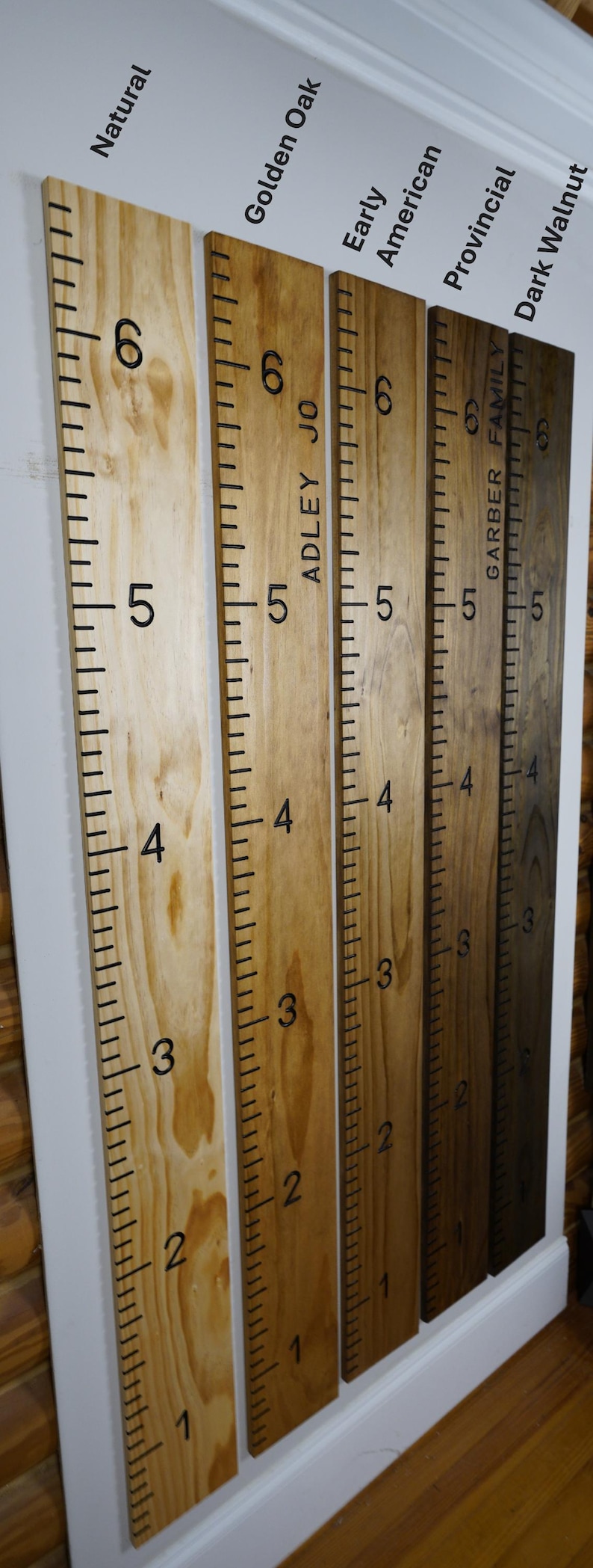 May include: Wooden growth charts in various finishes: Natural, Golden Oak, Early American, Provincial, and Dark Walnut. Each chart is marked with black numbers and lines, measuring up to 6 feet. Some charts include names.