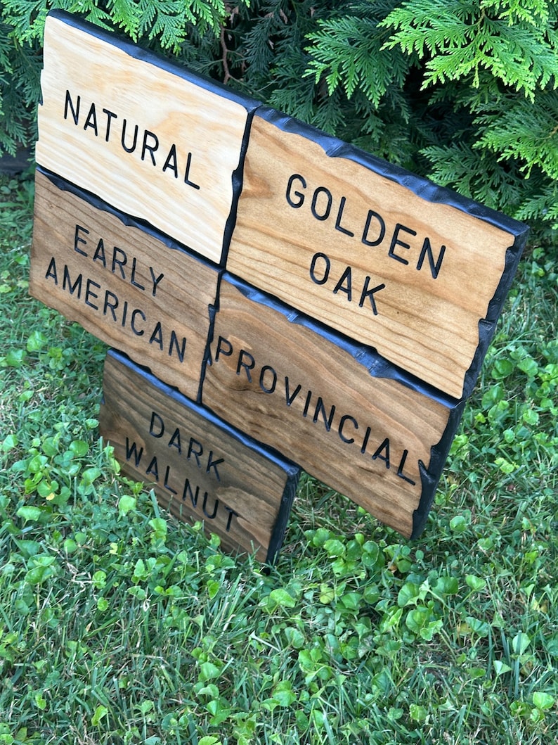 May include: A wooden sign featuring five different wood stain colors: Natural, Golden Oak, Early American, Provincial, and Dark Walnut. The sign is displayed outdoors on a bed of grass, with green foliage in the background.