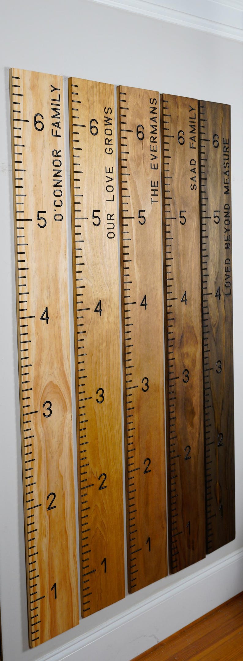May include: Wooden growth charts with black markings and family names. The charts are in varying shades of brown, with measurements in inches. The text includes "O'Connor Family", "Our Love Grows", "The Evermans", "Saad Family", and "Loved Beyond Measure".
