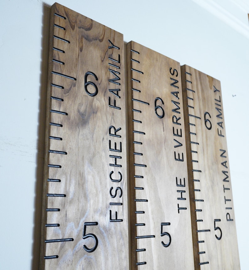 May include: Three wooden growth charts with black measurements and family names: Fischer Family, The Evermans, and Pittman Family. Each chart has numbers 5 and 6, with inch markings. The wood has a natural grain.