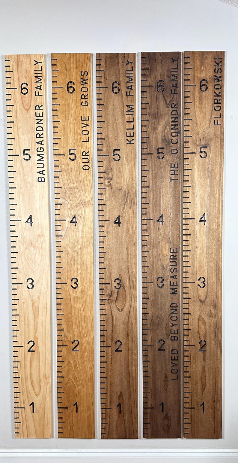 May include: Wooden growth charts with family names and measurements in inches. The charts are in various wood tones, with black markings and text. Each chart displays a family name and the phrase "Loved Beyond Measure."
