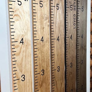 May include: Wooden growth charts with black markings and family names. The charts are in varying shades of brown and are marked with numbers and inch increments. The names "Sheahan Family", "The Mercados", "Teresi Family", "Loved Beyond Measure", and "Ayce Mckoy" are printed on the charts.