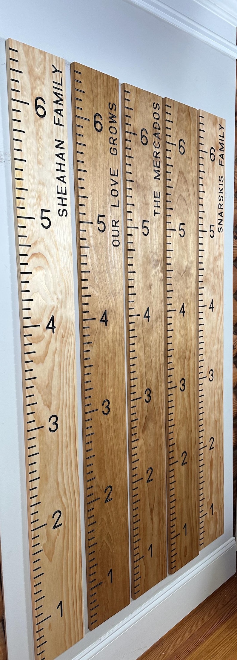 May include: Wooden growth chart rulers with black markings and family names. Each ruler is marked with numbers from 1 to 6, with family names like SHEAHAN FAMILY and SNARSKIS FAMILY. The rulers are mounted on a white wall.