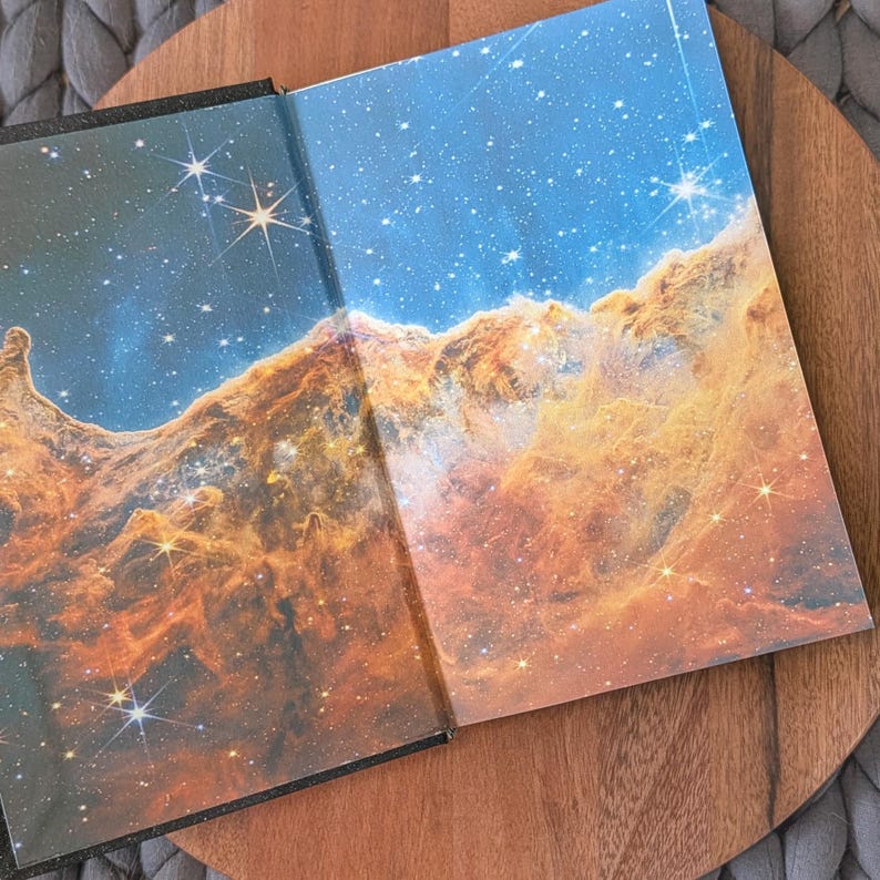 May include: An open hardcover book featuring a vibrant, high-resolution print of a nebula on the inside covers. The image displays swirling clouds of orange and gold cosmic dust set against a deep blue starry sky, with bright white starbursts.