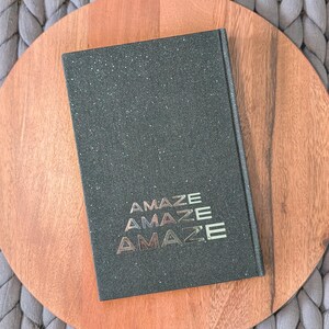 May include: A rectangular hardcover book with a dark green, glittery, textured cover. The word "AMAZE" is printed in a stacked, metallic, gold-toned font three times in the lower center of the front cover, creating a repeating visual effect.
