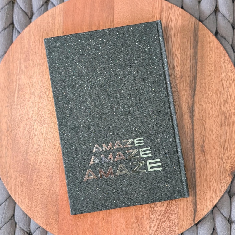 May include: A rectangular hardcover book with a dark green, glittery, textured cover. The word "AMAZE" is printed in a stacked, metallic, gold-toned font three times in the lower center of the front cover, creating a repeating visual effect.