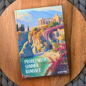 Może przedstawiać: Książka w twardej oprawie zatytułowana "Problematic Summer Romance" autorstwa Ali Hazelwood. Okładka przedstawia malowniczą ilustrację nadmorskiego miasteczka z kolorowymi budynkami, klifami i żywymi różowymi kwiatami. Książka spoczywa na drewnianej powierzchni.