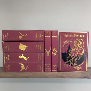 May include: A set of seven hardback books with burgundy covers and gold lettering. The books are the Harry Potter series by J.K. Rowling. The books are arranged in order of publication, with the first book, "Harry Potter and the Philosopher's Stone", on the right and the seventh book, "Harry Potter and the Deathly Hallows", on the left.