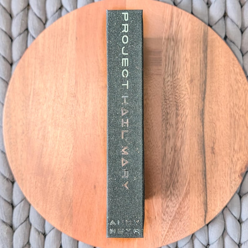 May include: A rectangular book spine with a dark, speckled charcoal gray finish. The text PROJECT HAIL MARY is printed vertically in a clean, light-colored sans-serif font, followed by the author name ANDY WEIR at the bottom of the spine.