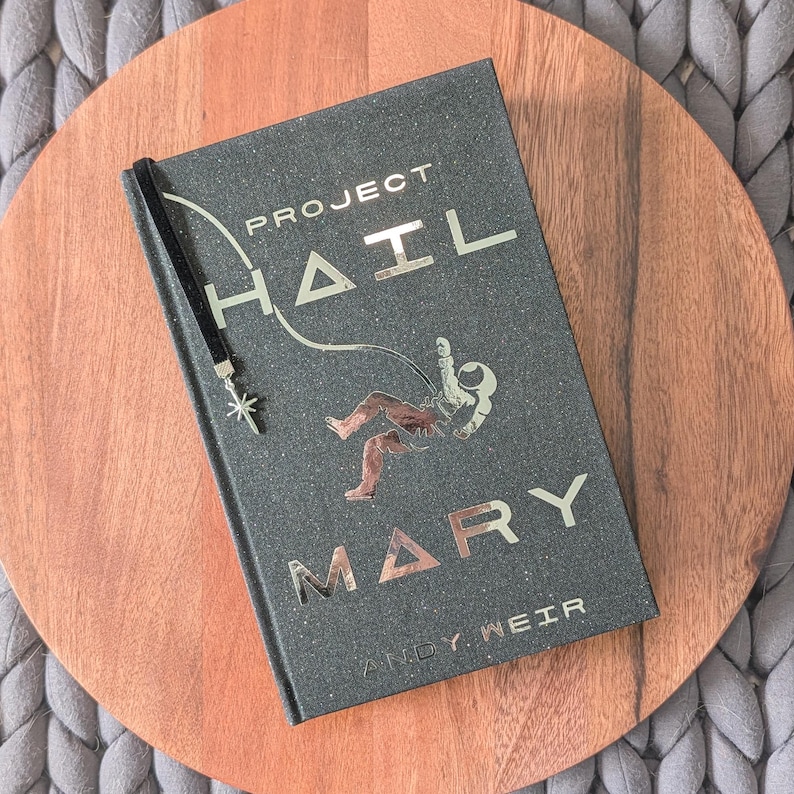 May include: A hardcover book titled Project Hail Mary by Andy Weir with a dark, speckled cover featuring metallic gold lettering and a graphic of an astronaut floating in space. A black velvet ribbon bookmark with a silver star charm is attached.