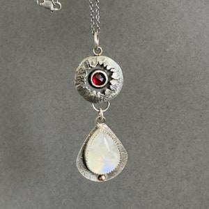 May include: A silver necklace with a teardrop-shaped moonstone pendant and a round pendant with a red garnet gemstone.