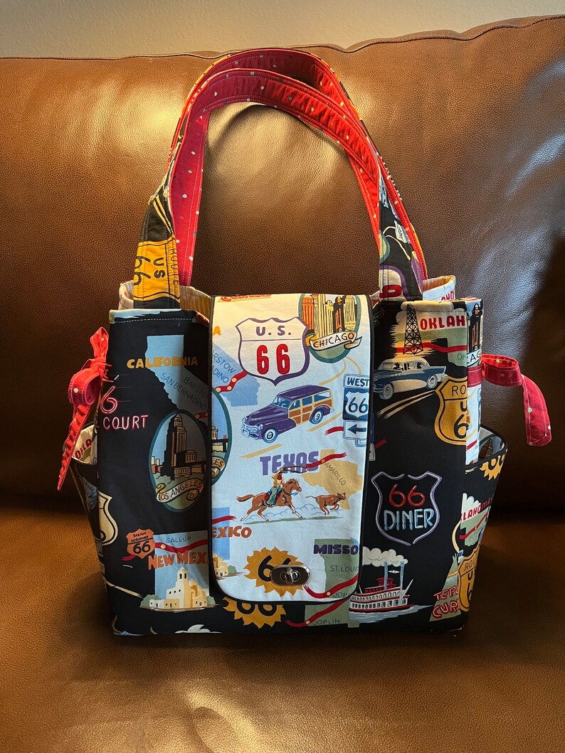 May include: A black tote bag with a red and white polka dot handle. The bag is decorated with a map of Route 66 and various landmarks along the route. The bag has a flap closure with a metal clasp.